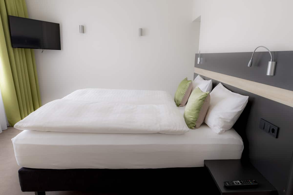 A modern hotel room with a neatly made bed, a flat-screen TV mounted on the wall, and a bedside table with a remote control.