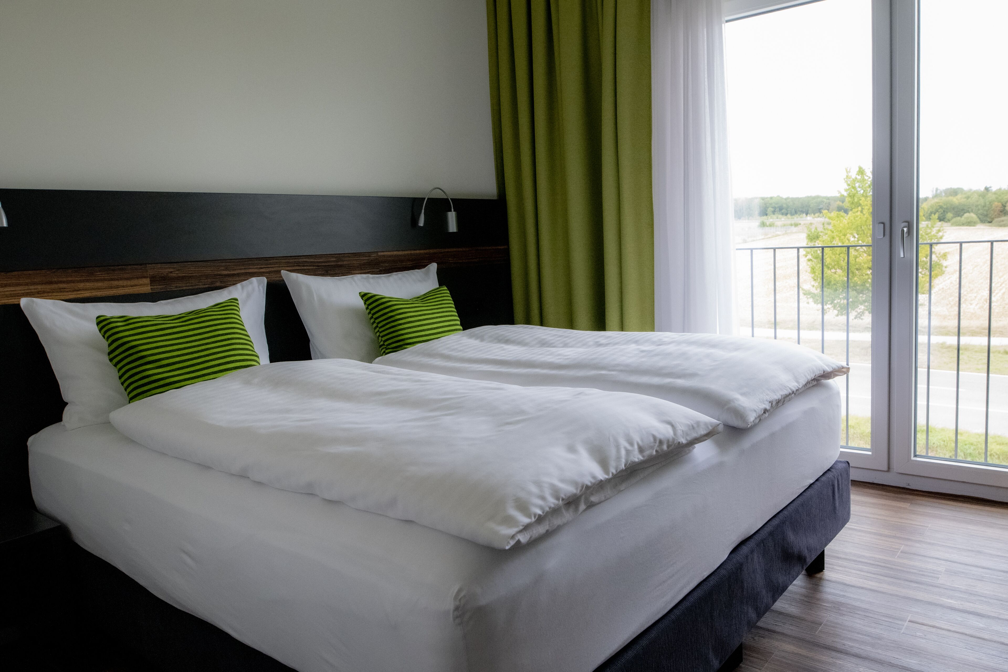 A double bed with white bedding and green striped pillows, a wooden headboard, a nightstand with a lamp, and a view of a balcony with a railing.