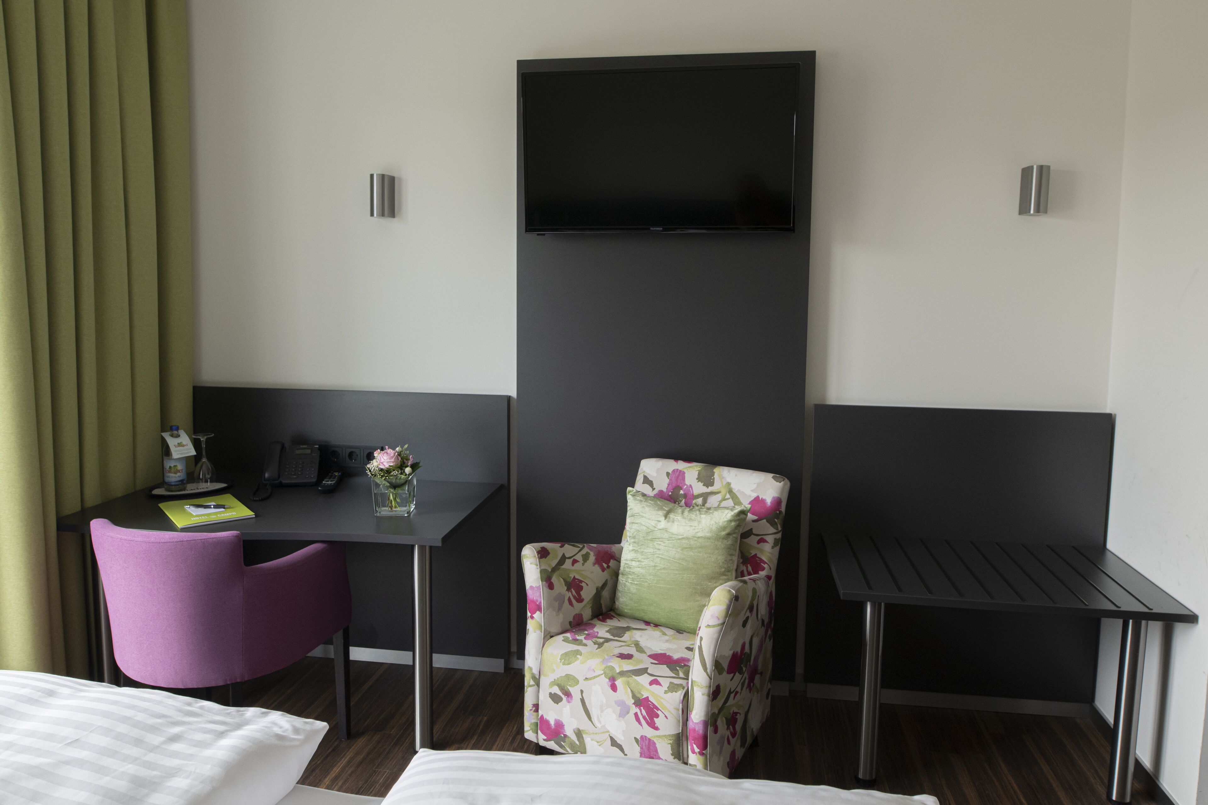 A hotel room with a bed, a desk with a chair, a television mounted on the wall, and a small table.