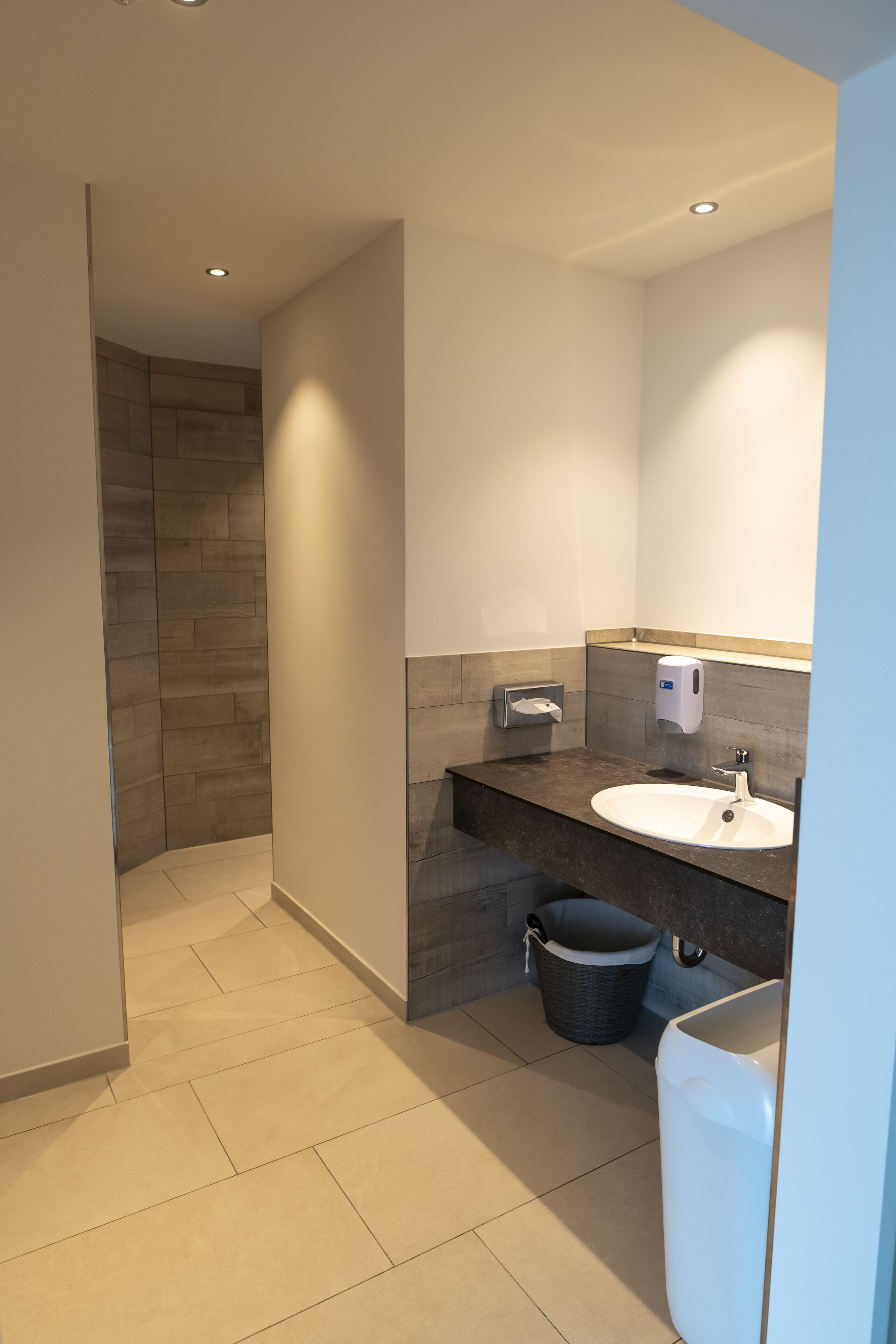 A modern bathroom with a sink, mirror, towel dispenser, and a trash bin.