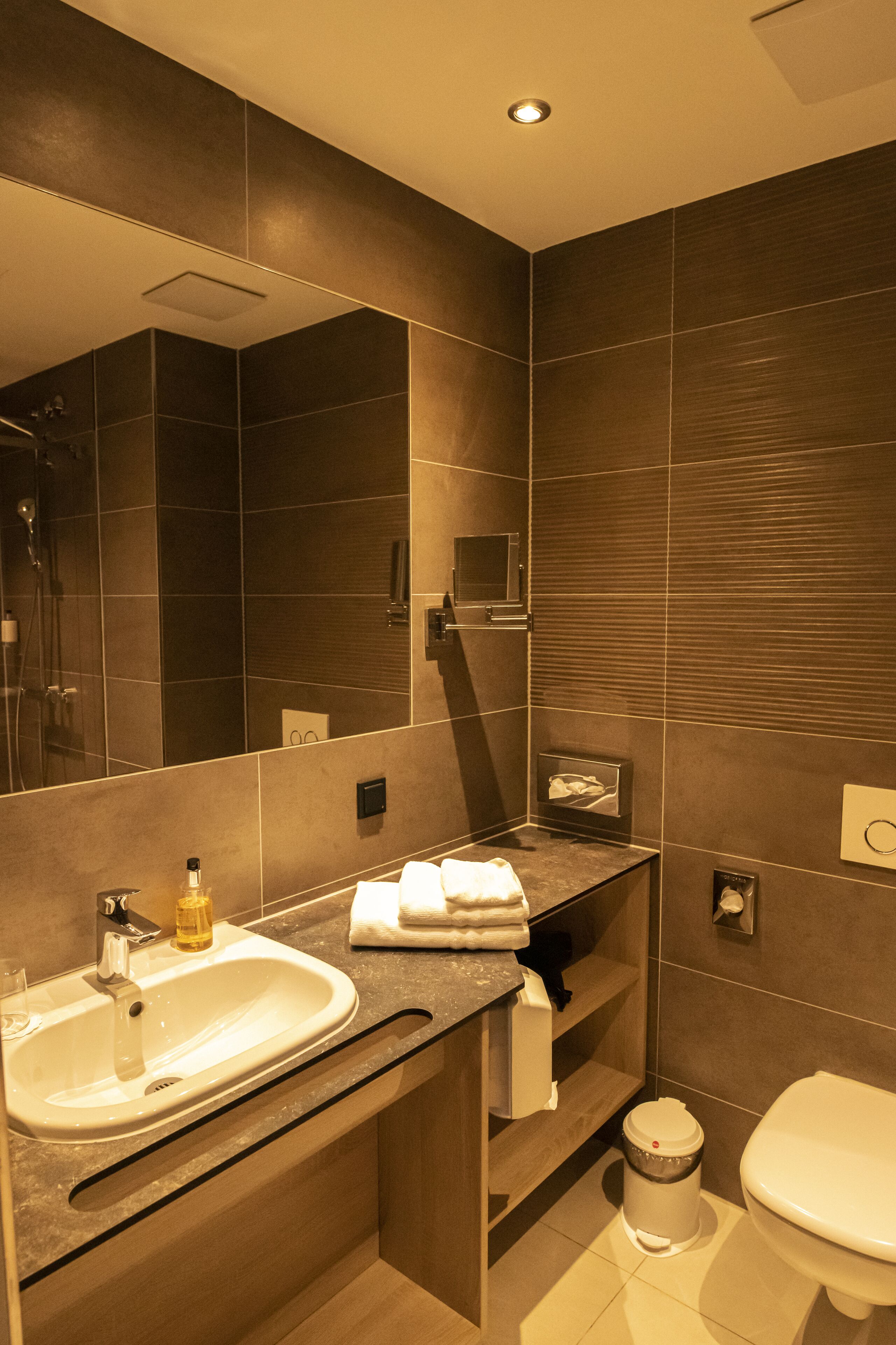 A modern bathroom with a large mirror, a sink, a toilet, and a trash can.