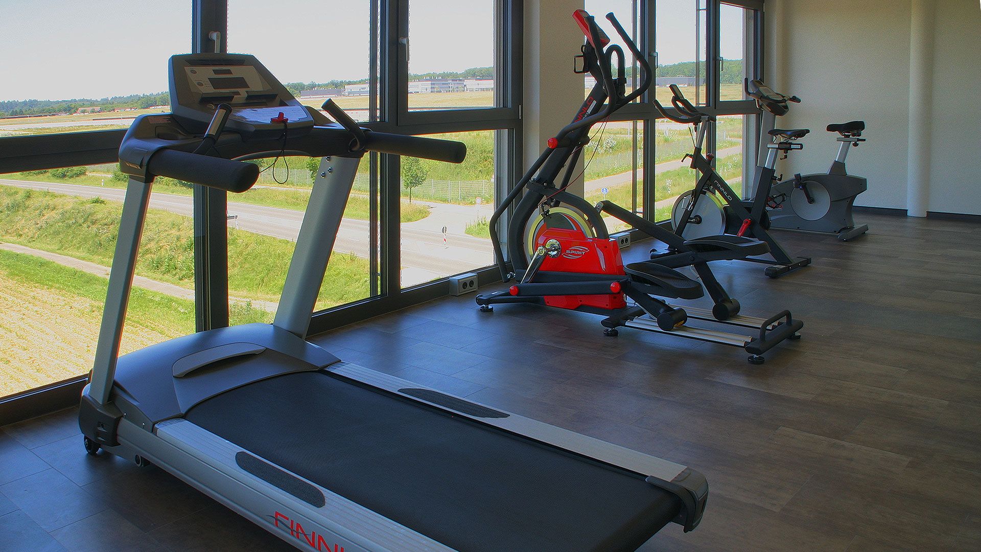 Ett gym med löpband, motionscyklar och en roddmaskin, stora fönster med utsikt över en väg och grönområden.