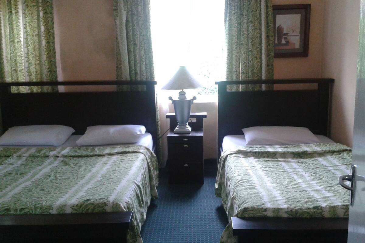A hotel room with two single beds, a nightstand with a lamp, and a window with curtains.