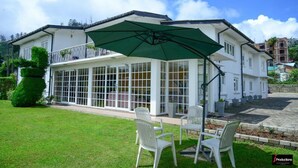 A white house with a large glass conservatory, outdoor seating, and a green umbrella. - Country House - Nuwara Eliya (Nuwara Eliya)