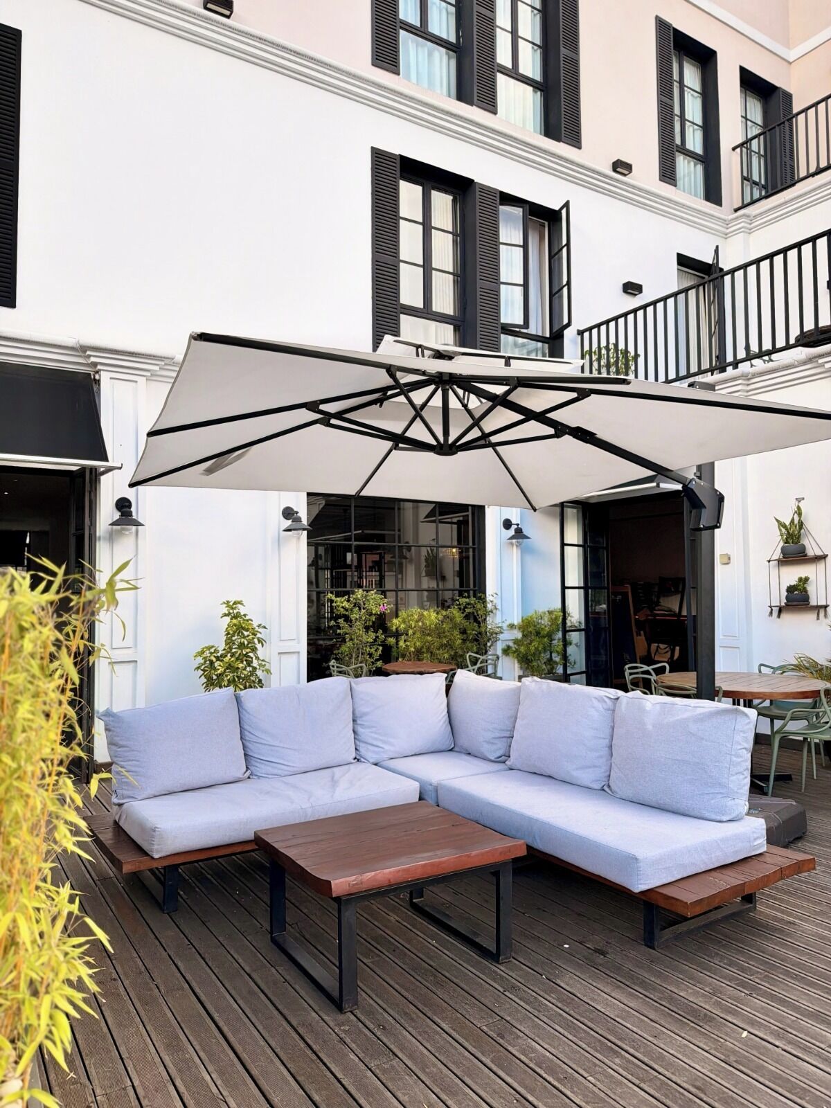 Terraza de madera con sofá seccional y una mesa, rodeada de plantas en macetas y un toldo blanco.