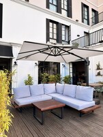 Terraza de madera con sofá seccional y una mesa, rodeada de plantas en macetas y un toldo blanco.