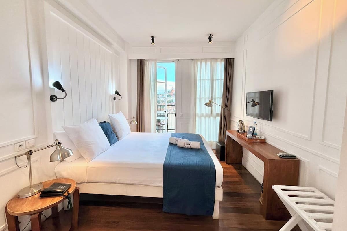 A hotel room with a bed, bedside table, lamp, TV, and a view of the city.
