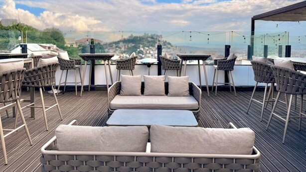 Terraço ao ar livre com mobiliário de vime, incluindo um sofá e mesas, com vista para a cidade.