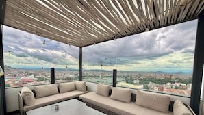Uma área de estar coberta ao ar livre, com um sofá e uma mesa, e amplas janelas com vista para a cidade.
