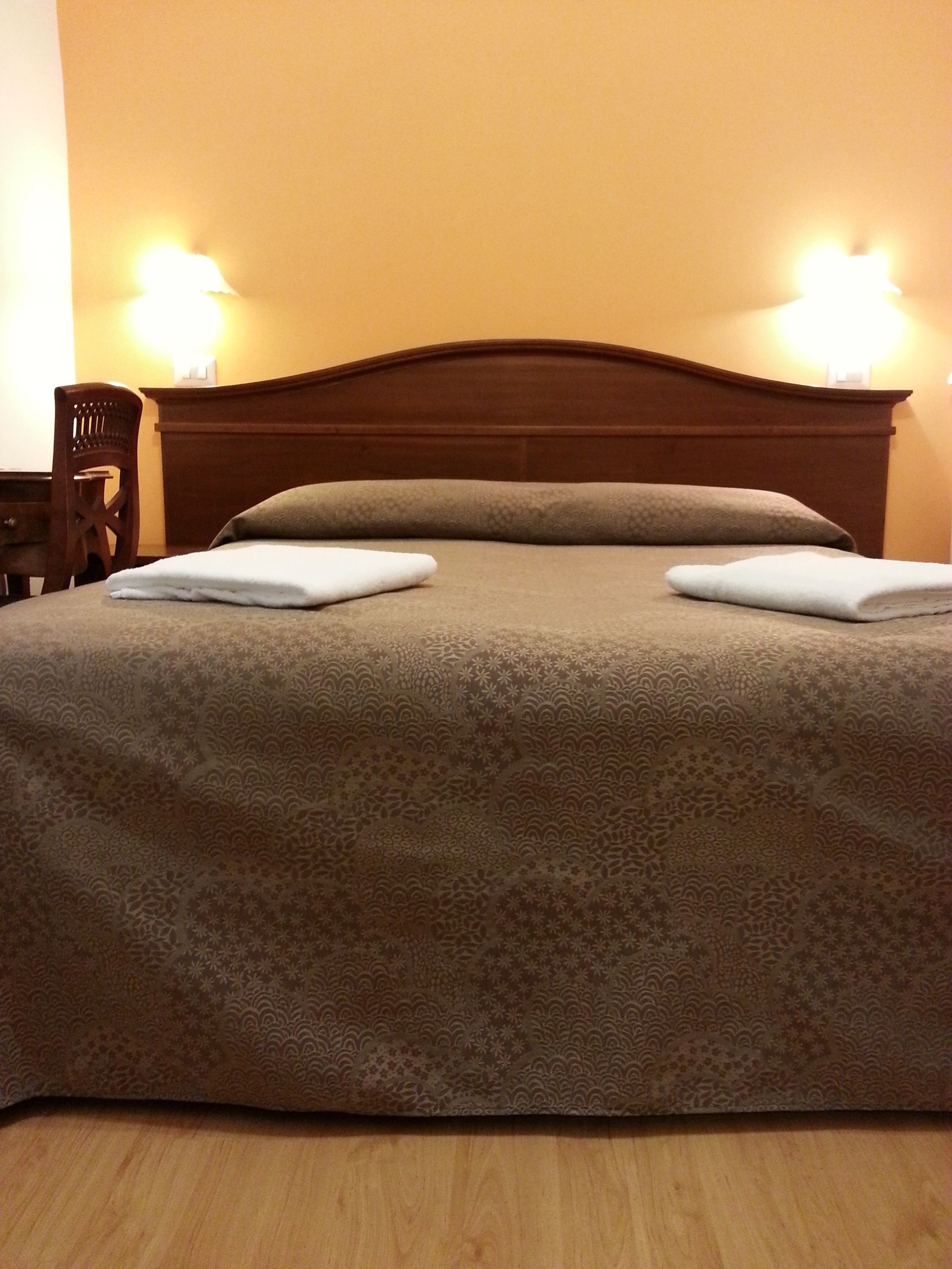 A neatly made bed with a patterned bedspread and two pillows, a wooden nightstand with a lamp, and a chair in a room with a wooden headboard.