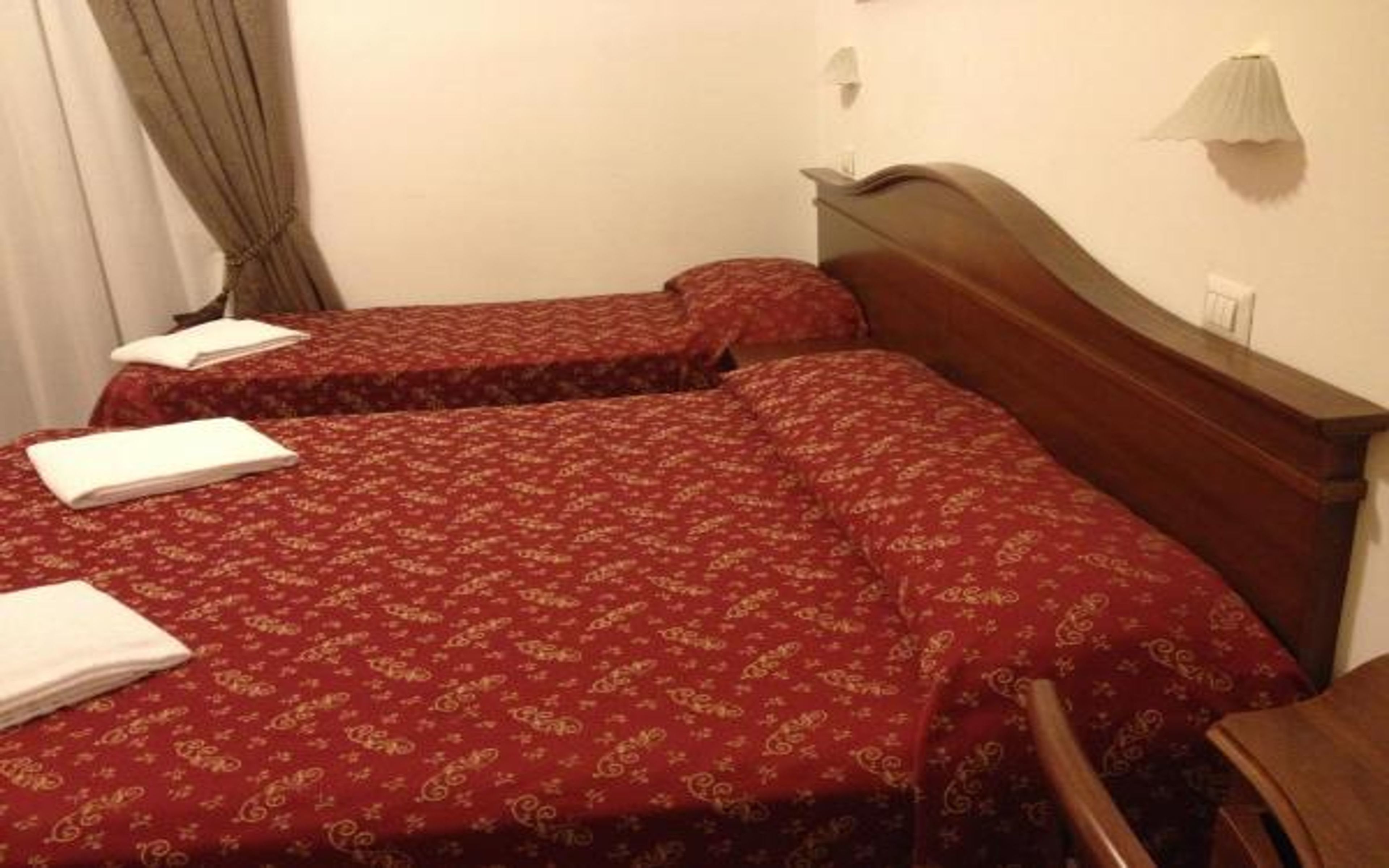 A hotel room with two beds, each with a red patterned bedspread and white pillows. There is a wooden headboard and a small table with a lamp.