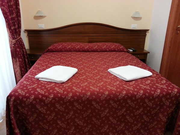 Una cama bien hecha con una colcha roja estampada y dos toallas blancas dobladas.