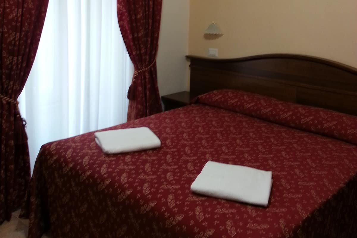 A hotel room with a bed, red curtains, a nightstand, and a lamp.