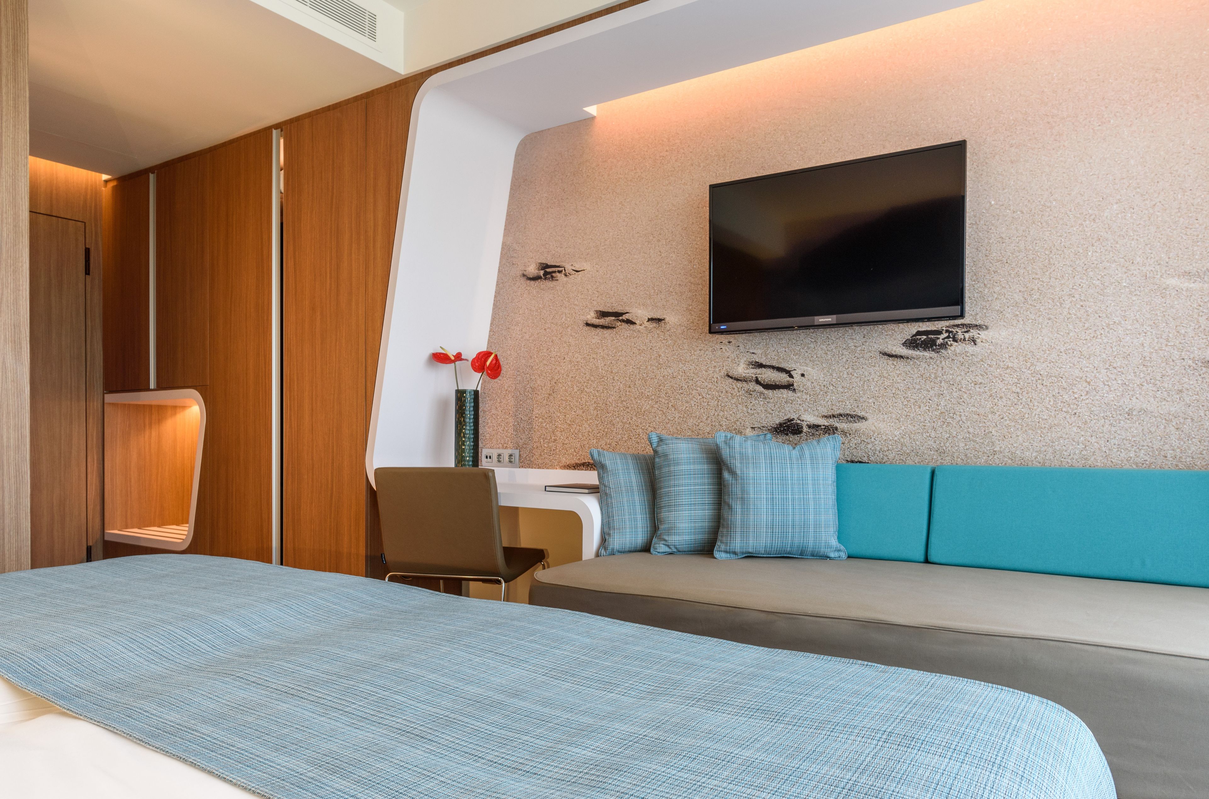 A modern hotel room with a bed, a sofa, a TV mounted on the wall, and a desk with a chair.