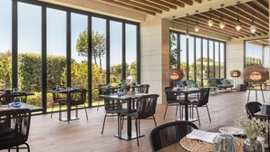 Un amplio espacio exterior con zona de comedor, varias mesas y sillas, ventanas grandes con vistas a la vegetación y lámparas colgantes.