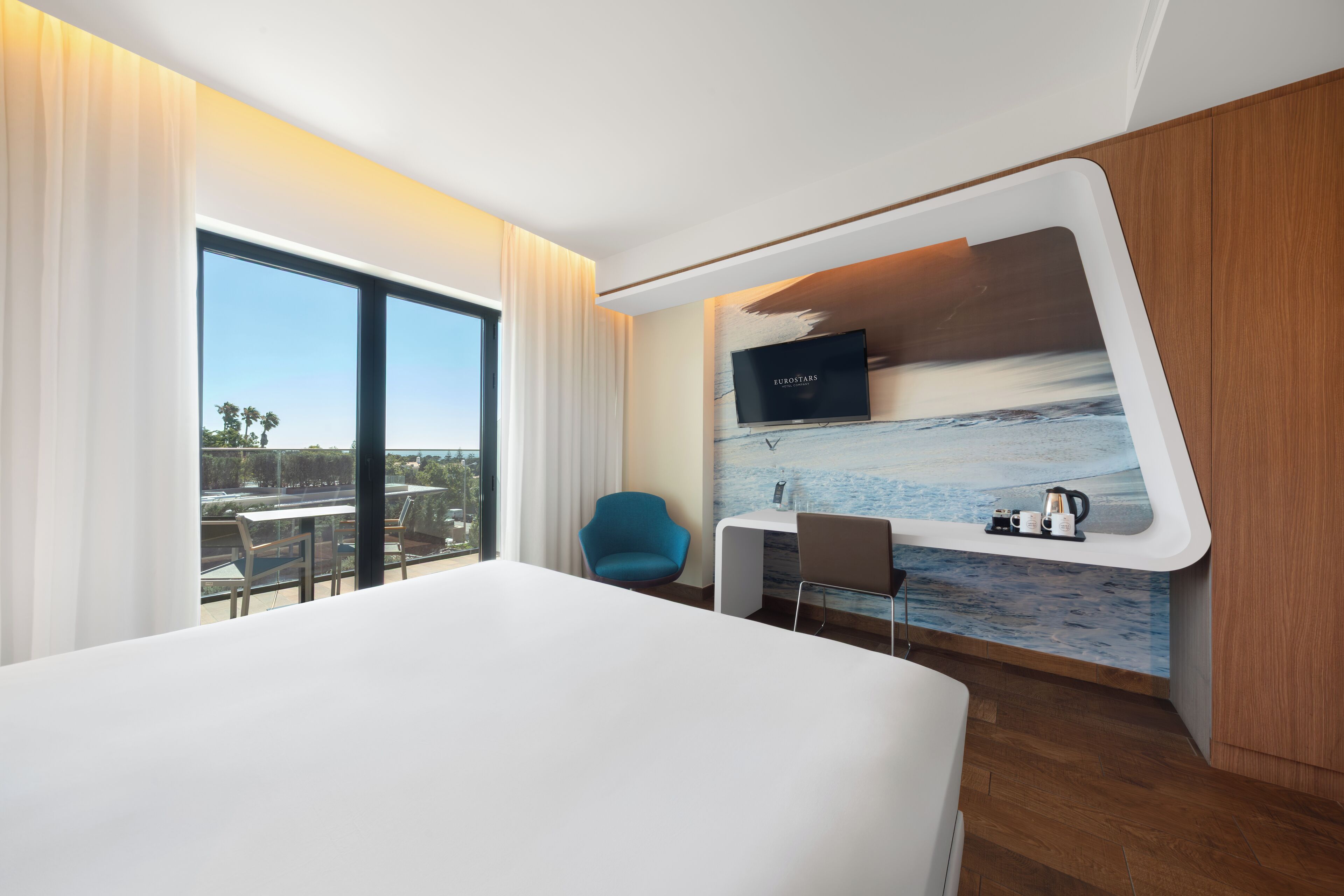 Ein modernes Hotelzimmer mit einem großen Bett, einem Schreibtisch, einem Sessel und einem an der Wand befestigten Fernseher.