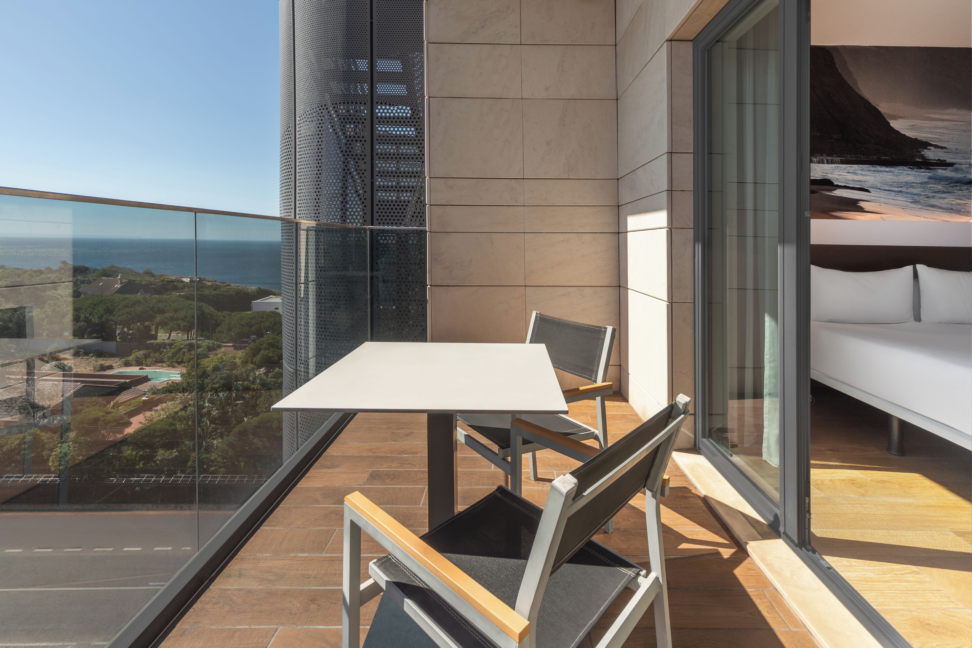 Een balkon met een tafel en stoelen, een witte bank en een groot raam met uitzicht op zee en gebouwen.