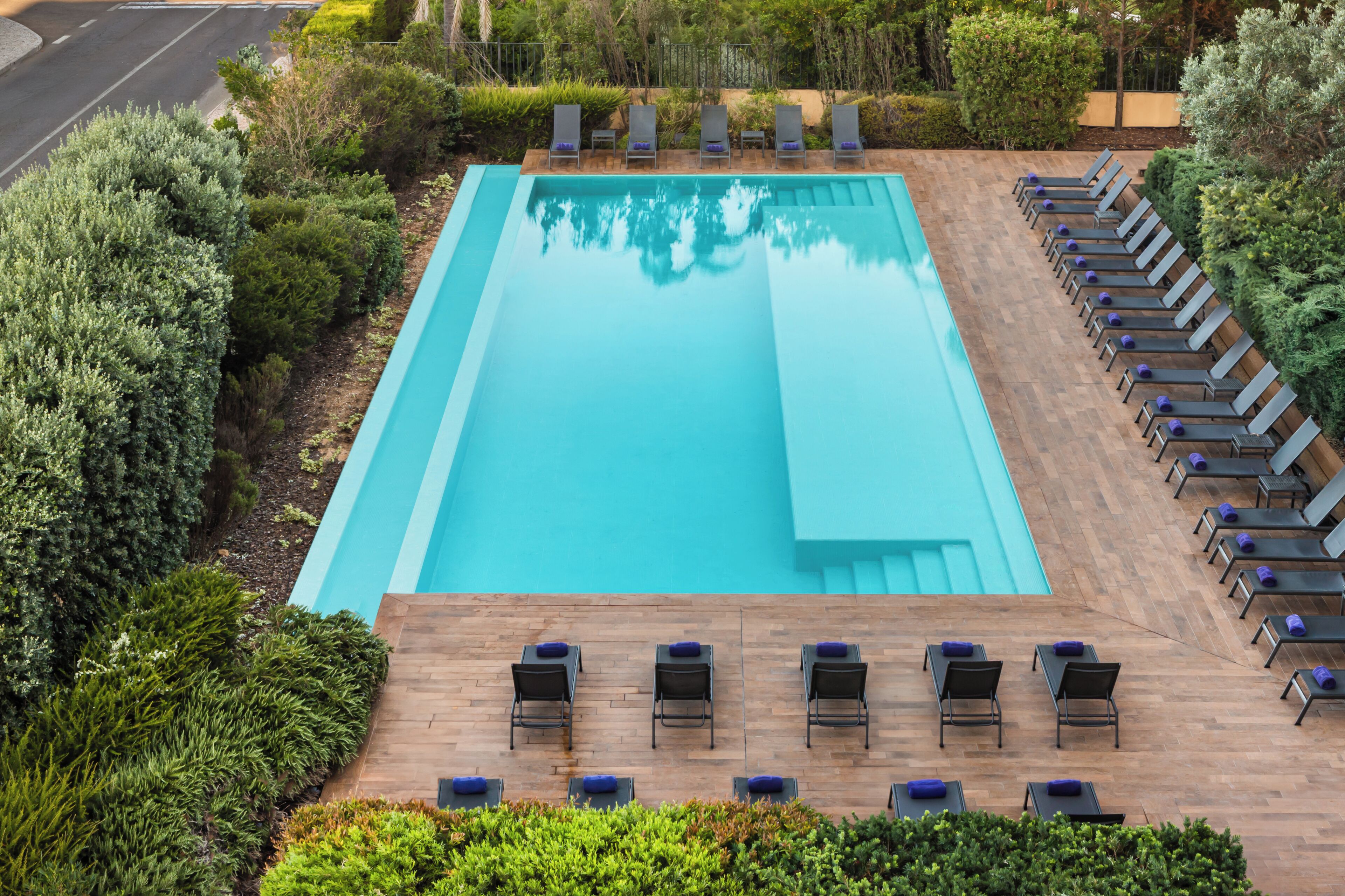 A rectangular swimming pool with a wooden deck surrounding it, equipped with lounge chairs and tables.