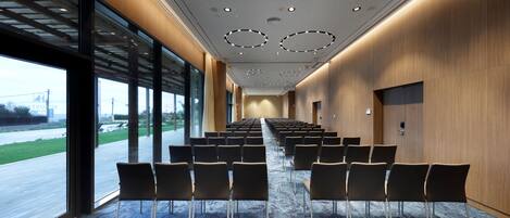 Un salón de conferencias con filas de sillas, ventanas amplias y una pared de madera.