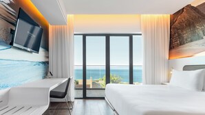 Un hotel moderno con una ventana grande que ofrece vista al mar, un televisor de pantalla plana en la pared y una cama con sábanas blancas.