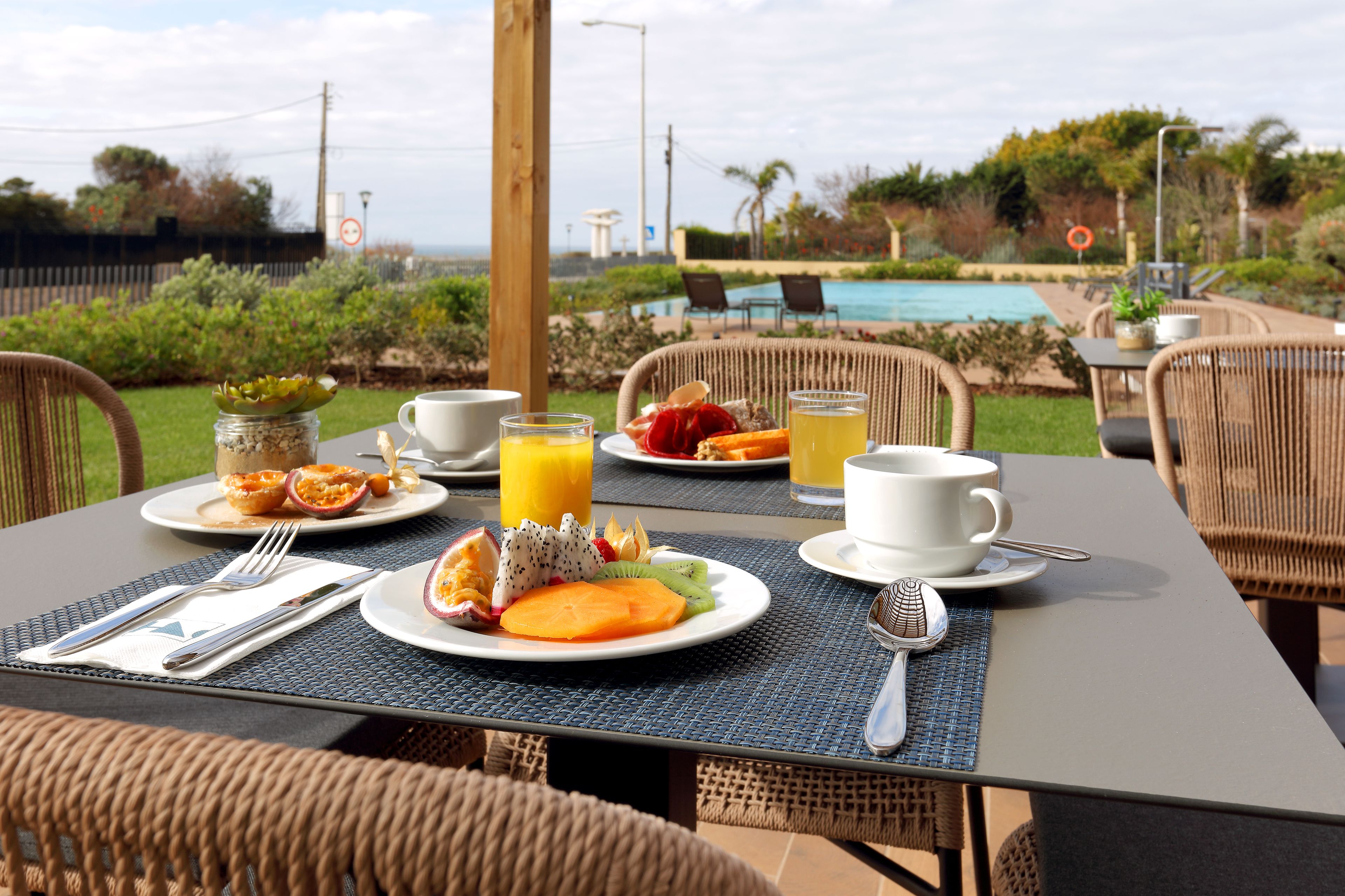 Una mesa lista para el desayuno, con platos de fruta, un vaso de zumo de naranja y una taza de café, con vistas a una zona de piscina.