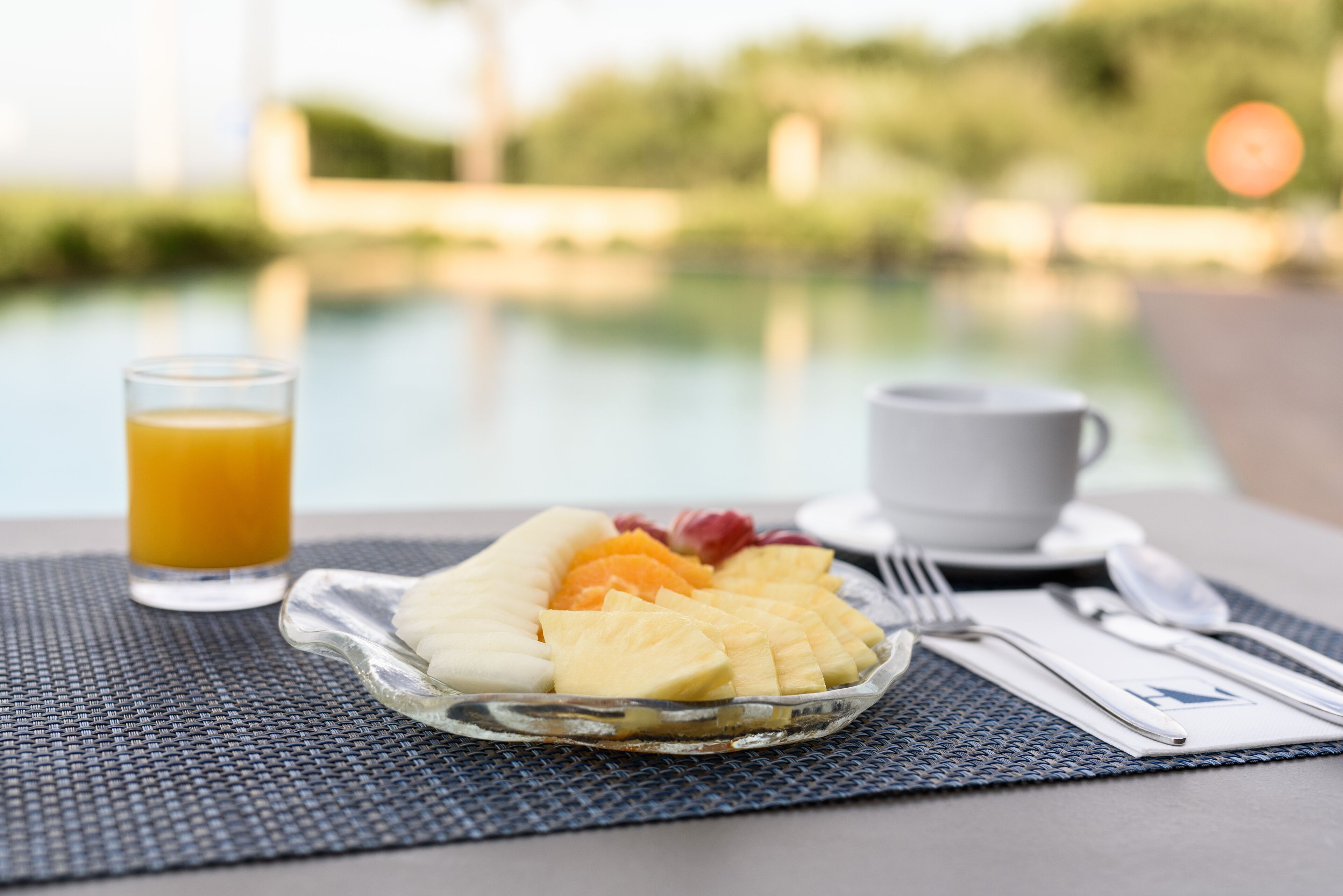 Un desayuno con un plato de fruta, un vaso de zumo de naranja y una taza sobre una mesa junto a una piscina.