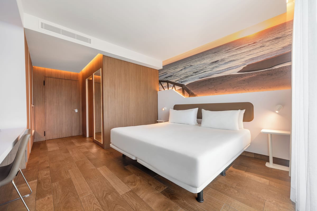 A modern hotel room with a large bed, wooden flooring, a desk, and a wall-mounted artwork.