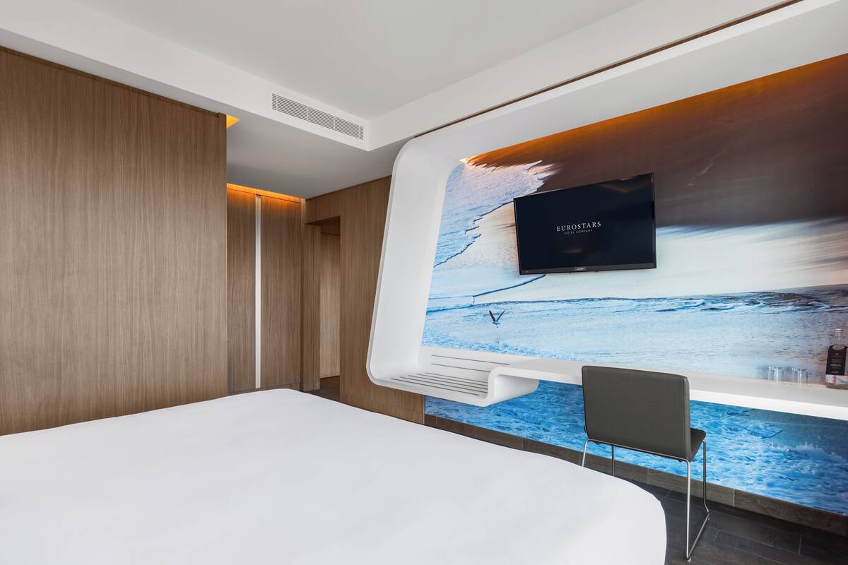 A modern hotel room with a large bed, a flat-screen TV mounted on a wall with a wave-themed wallpaper, and a small seating area with a chair.