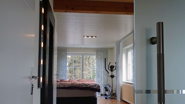 A bedroom with a wooden floor, a bed with a patterned bedspread, a standing exercise bike, and a window with a view of trees. - Hannover Guest House (Hannover)