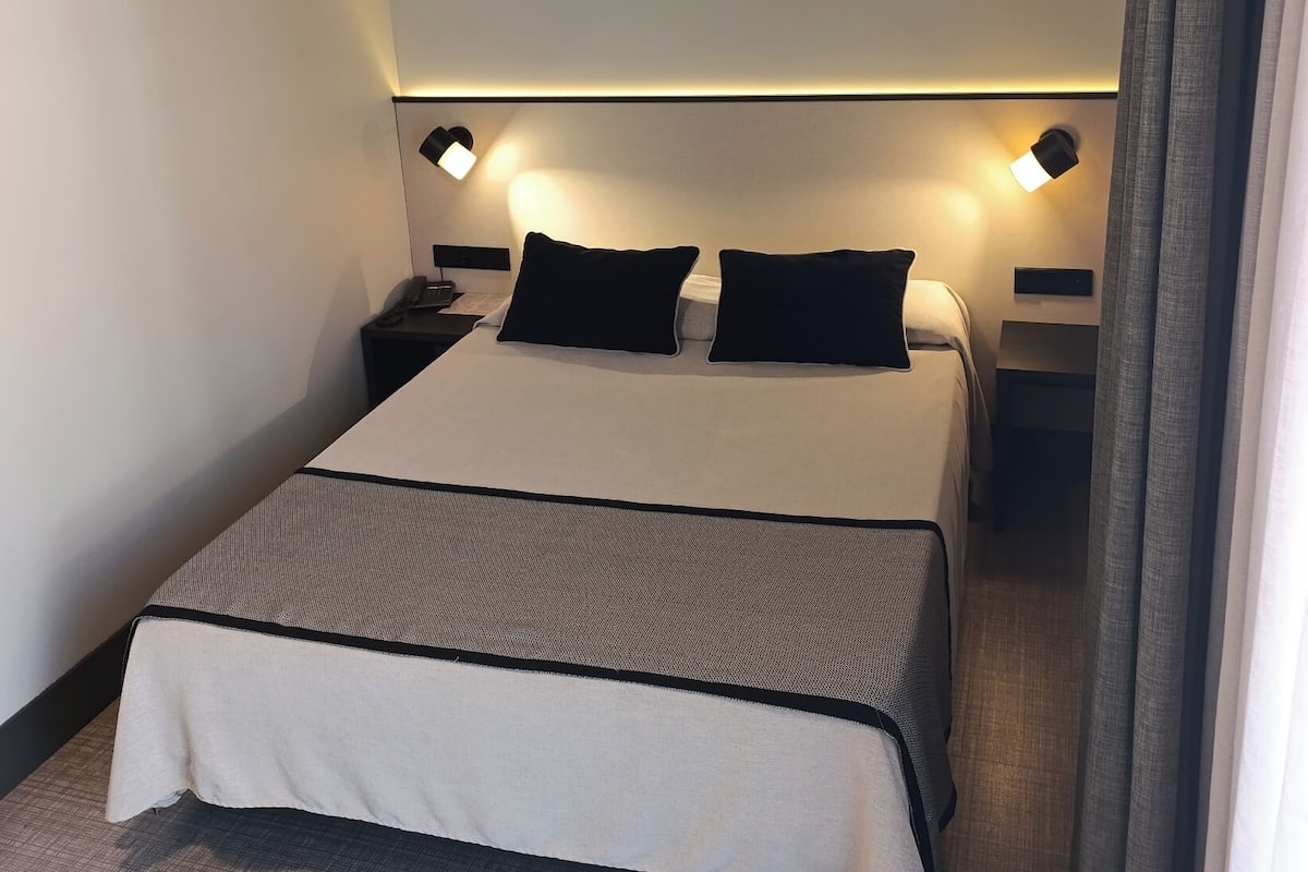 Una cama bien hecha con una colcha blanco y gris, dos almohadas negras y dos lámparas de noche a cada lado.