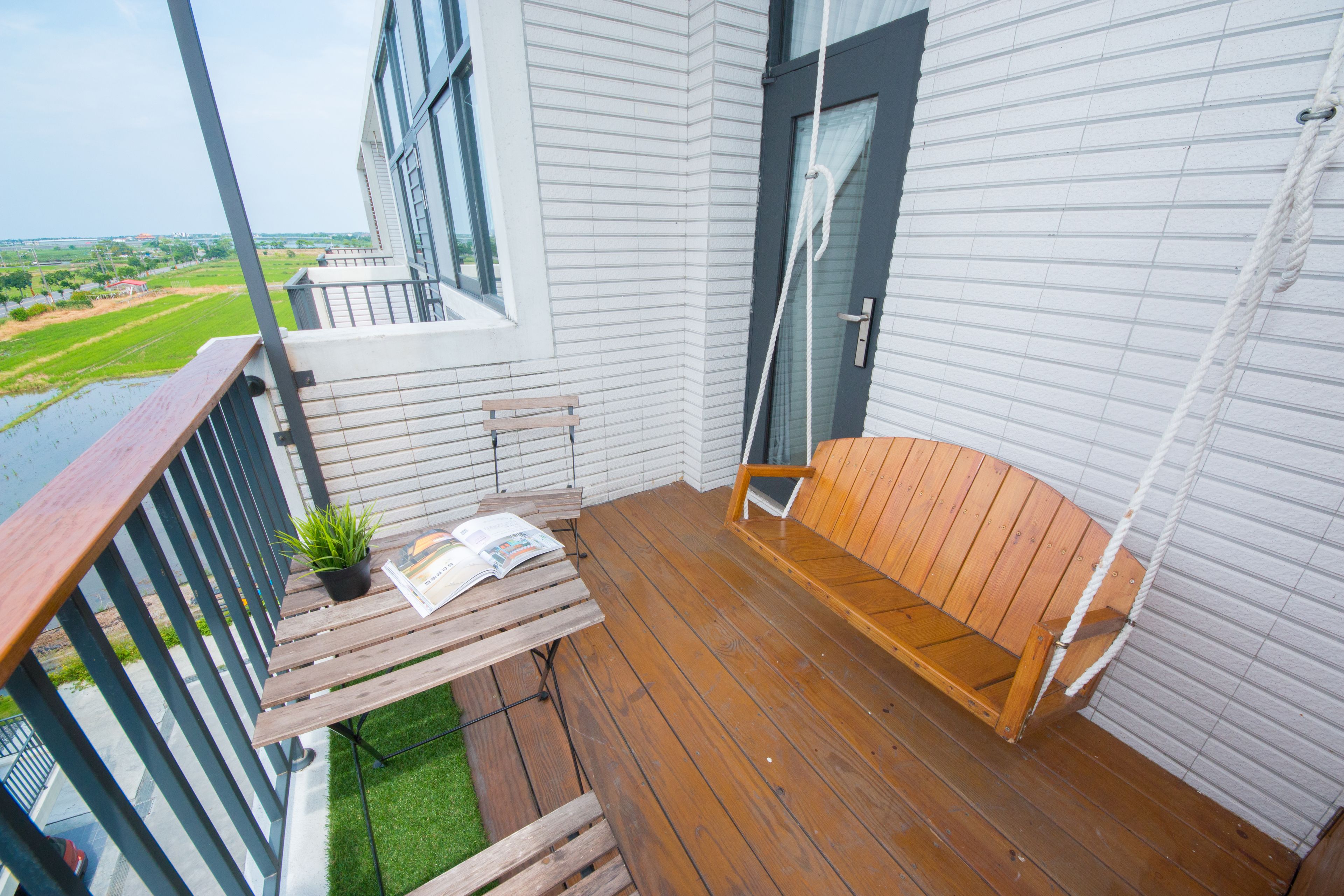 A balcony with a wooden bench, a small table with a plant and a magazine, and a view of a green field and distant buildings.