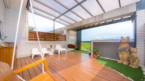 Une vaste terrasse dotée d’un plancher en bois, de chaises en plastique blanches, d’une balançoire en bois et offrant une vue sur la mer.