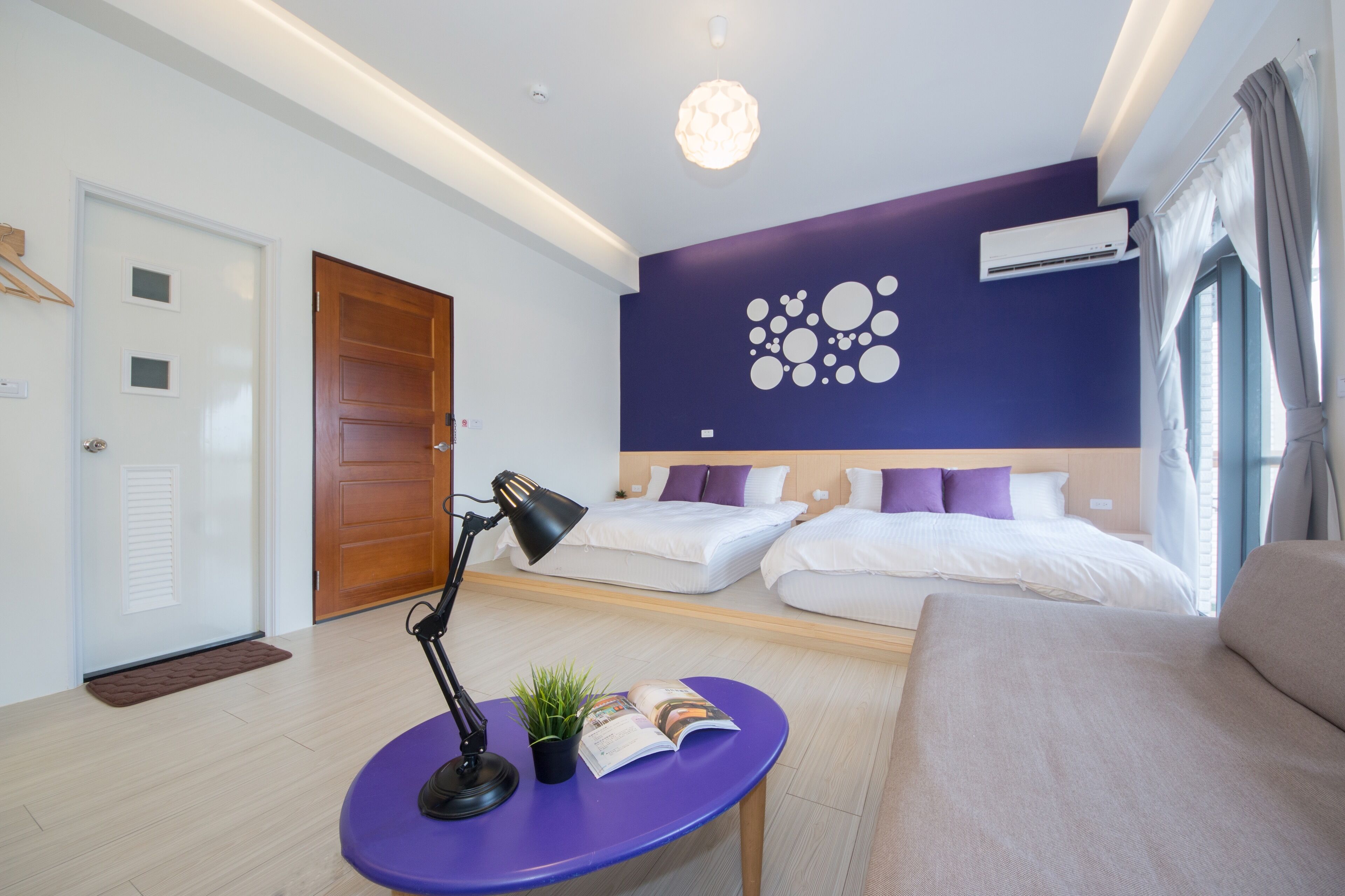 A modern hotel room with a wooden door, a purple table with a lamp and a plant, two beds with white and purple bedding, and a ceiling light.