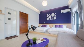 A modern hotel room with a wooden door, a purple table with a lamp and a plant, two beds with white and purple bedding, and a ceiling light.