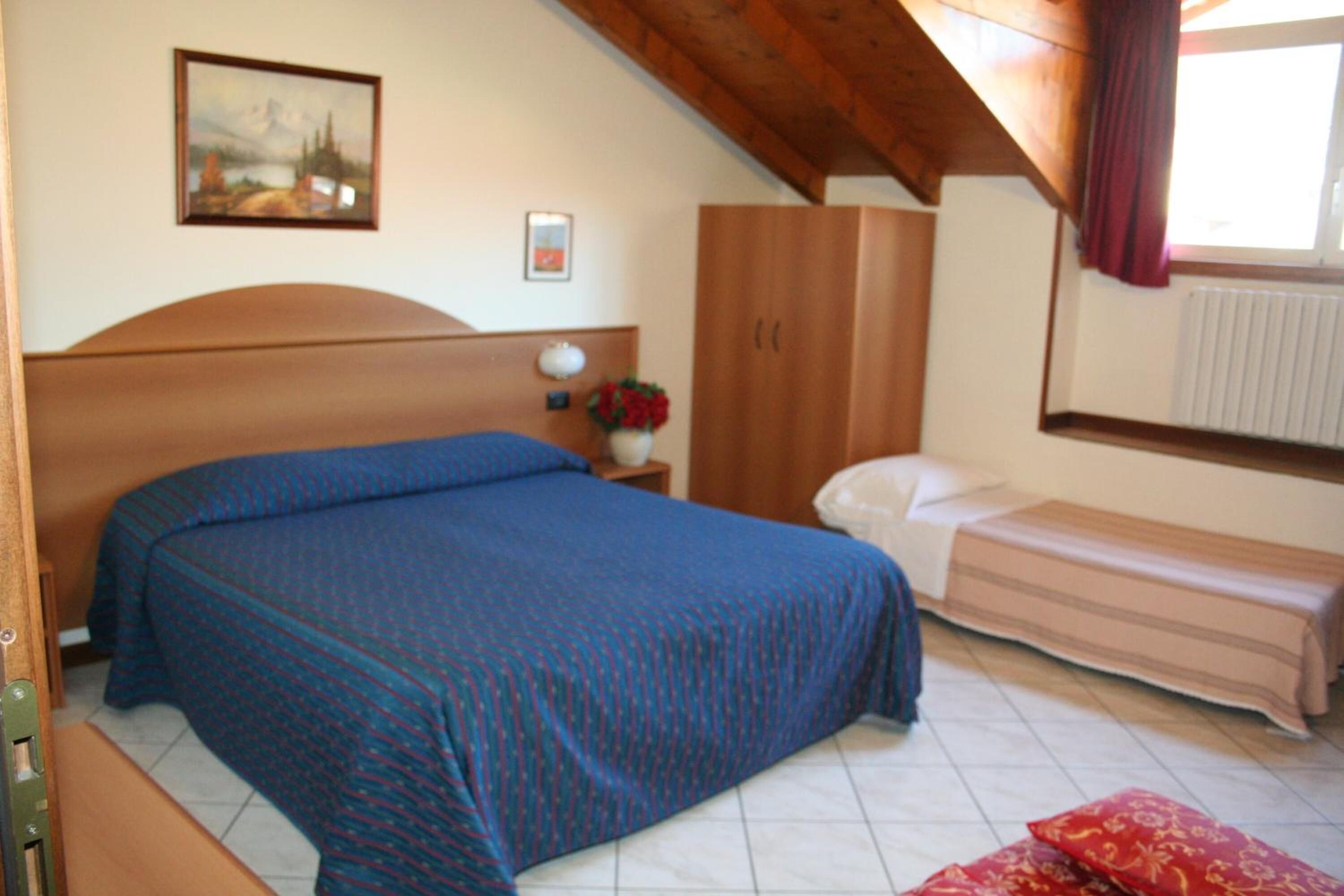 A bedroom with a large bed, a smaller bed, a wardrobe, a nightstand with a lamp, and a painting on the wall.