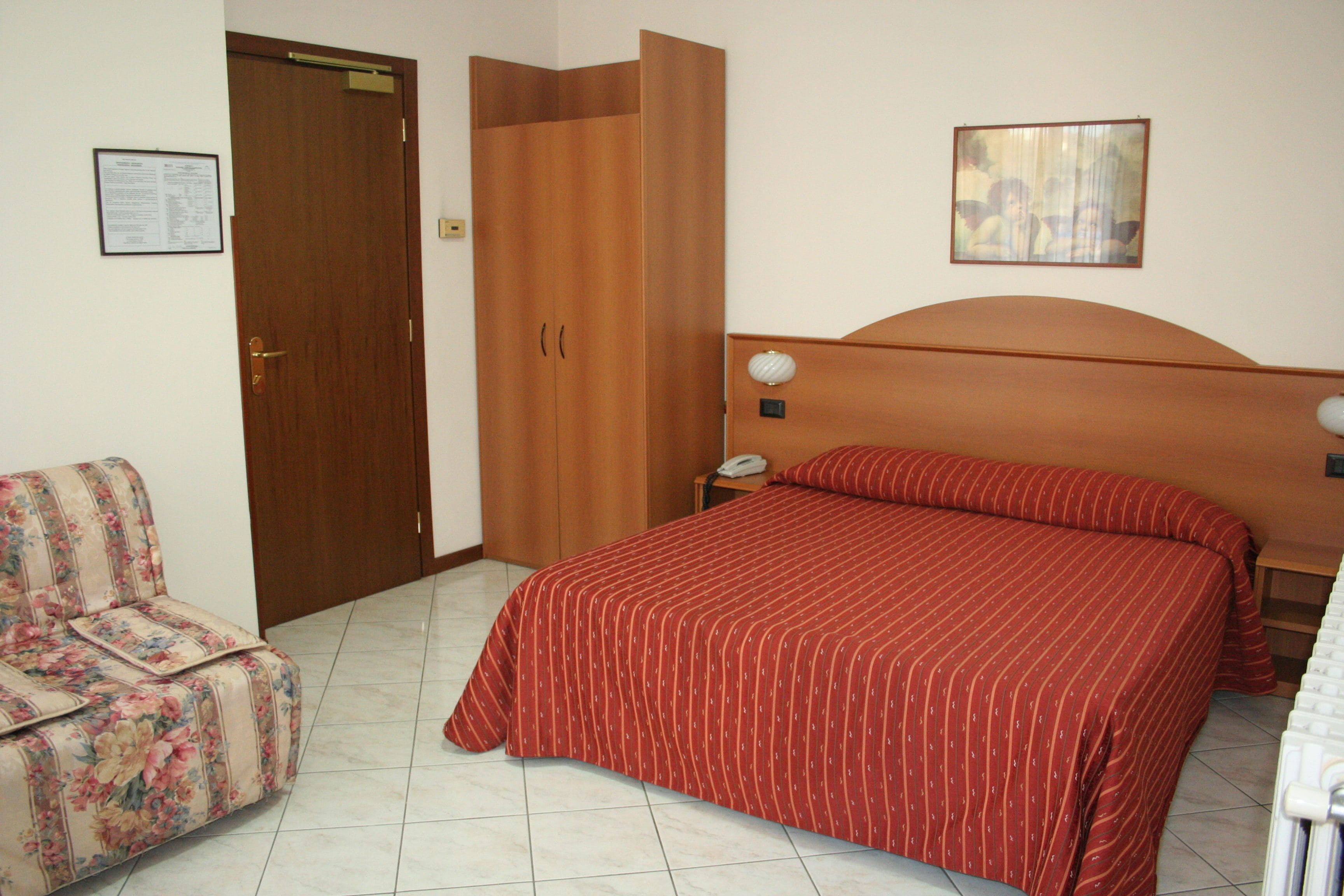 A bedroom with a bed, a wooden wardrobe, a floral armchair, a nightstand, and a wall-mounted lamp.