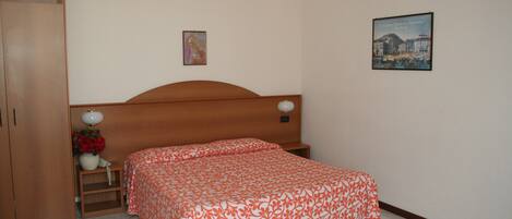 Un dormitorio con cabecera de madera, una cama individual con colcha estampada de flores naranjas, una mesita de noche con lámpara, una pequeña mesa con un florero y un cuadro en la pared.