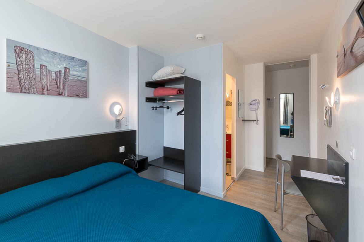 A modern hotel room with a large bed, a built-in wardrobe, a desk, and a bathroom visible through an open door.