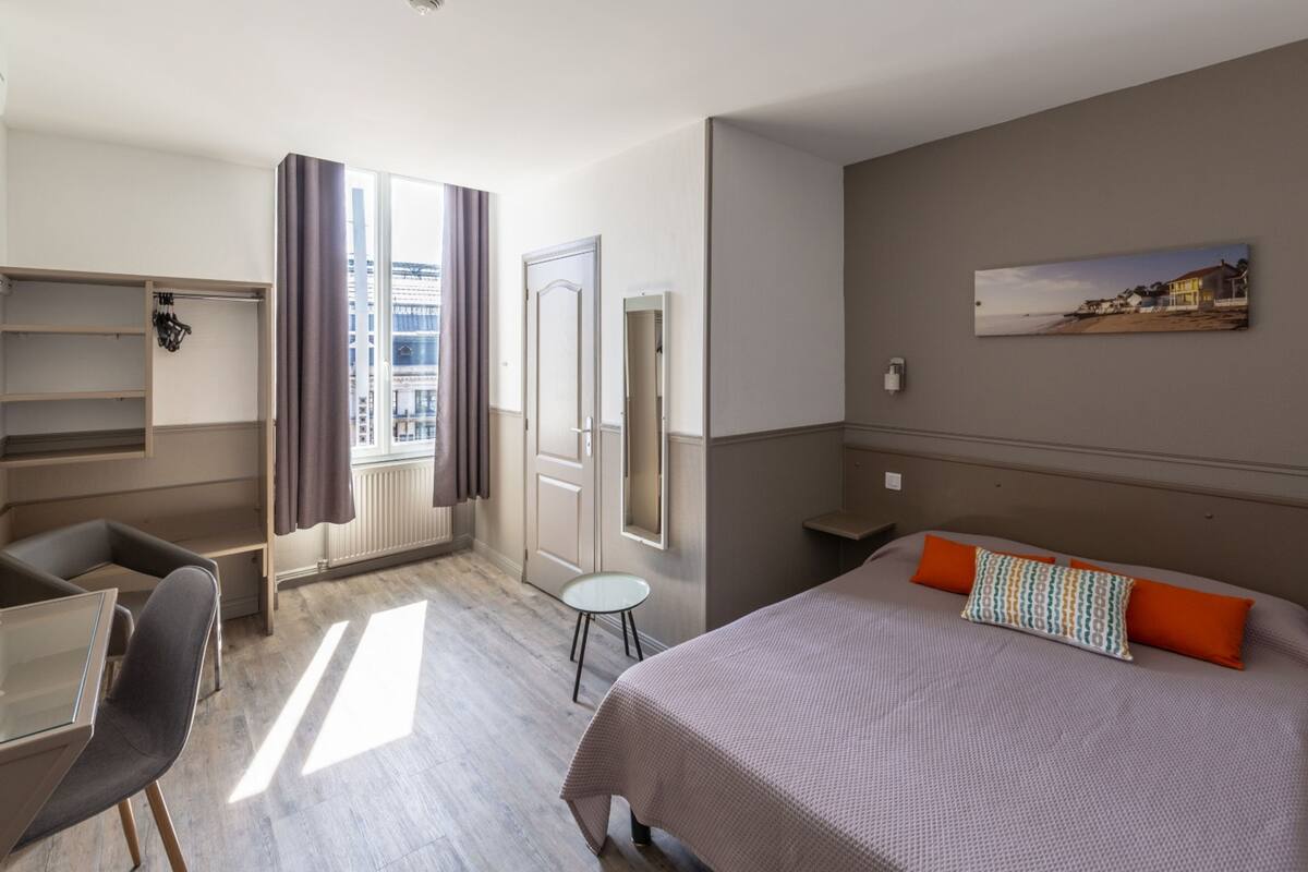 A hotel room with a bed, a desk, a chair, a small table, a wardrobe, a window with curtains, and a painting on the wall.