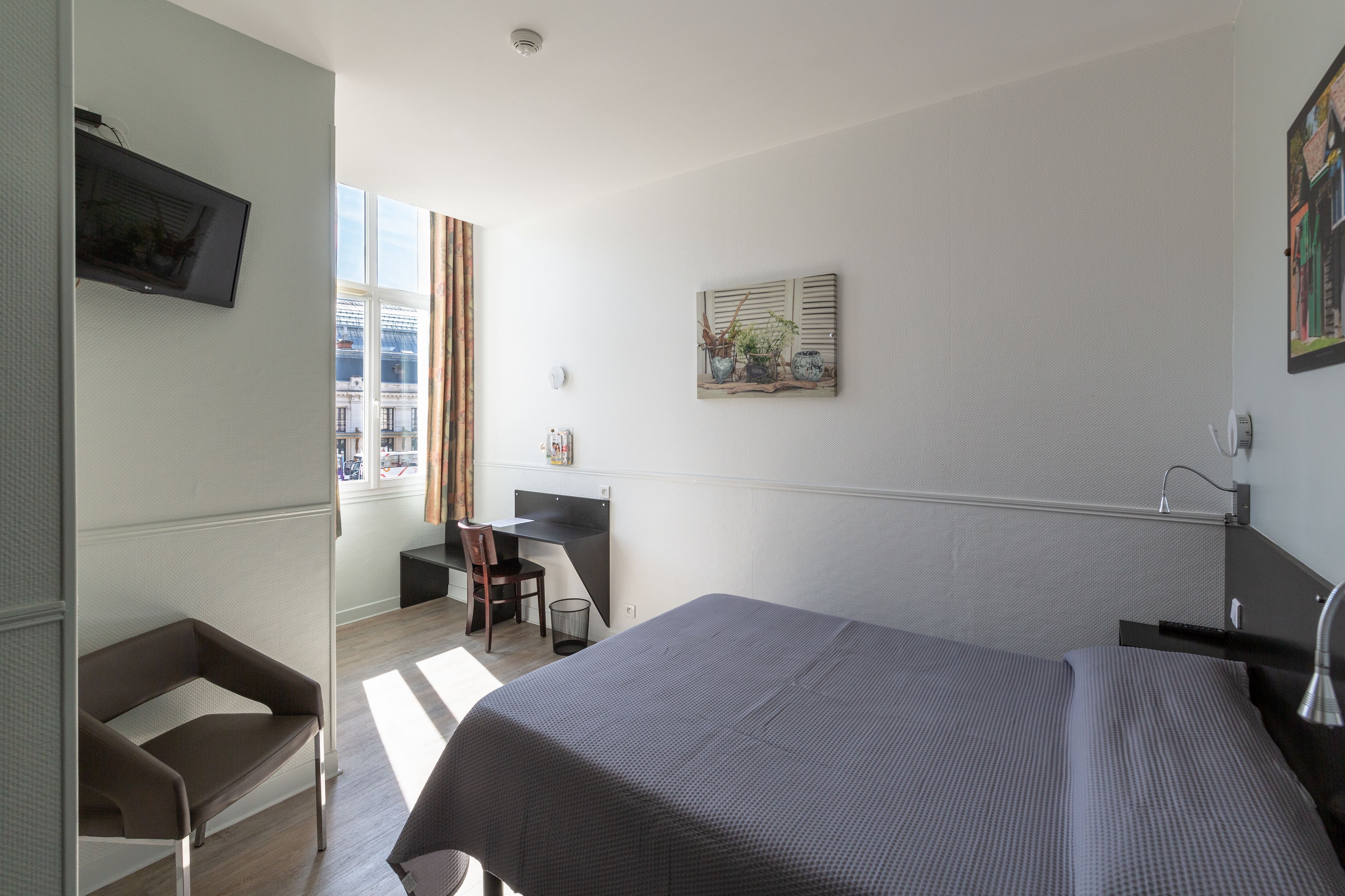 Une chambre d’hôtel avec un lit, un bureau et une chaise. Il y a une fenêtre avec des rideaux, un tableau au mur et un téléviseur fixé au mur.