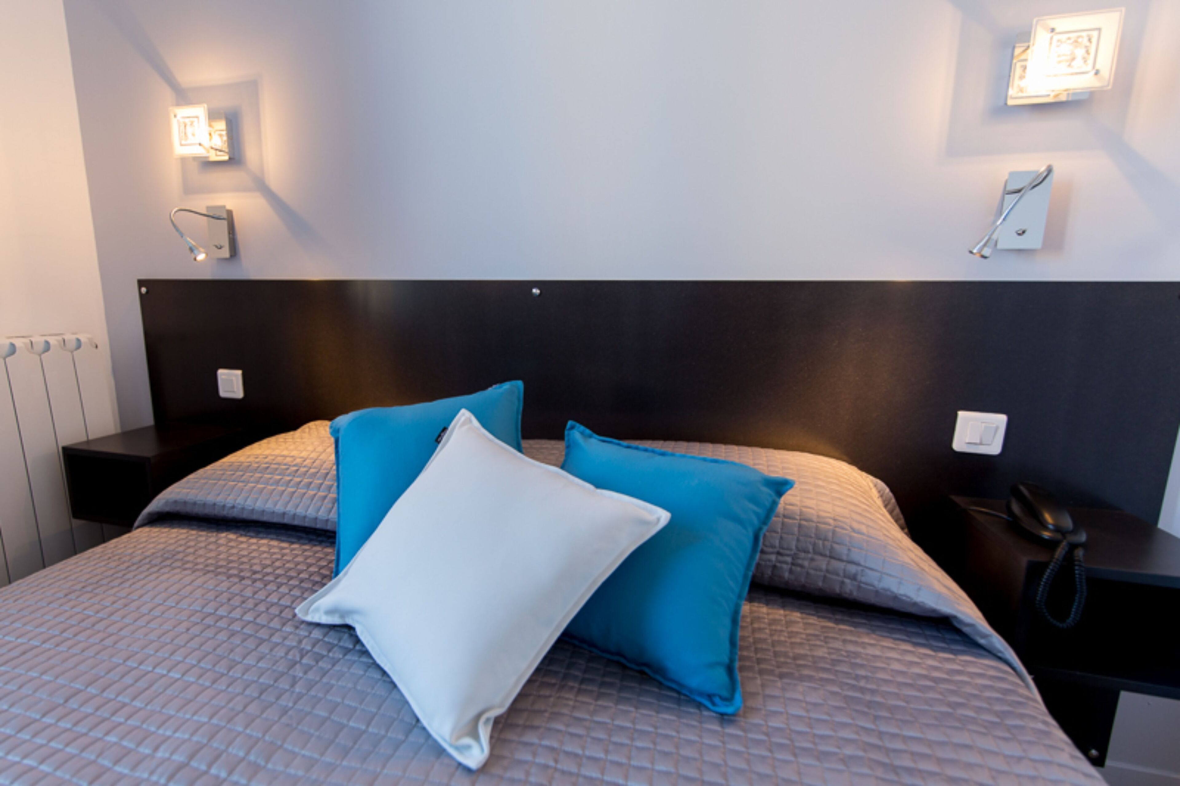 A neatly made bed with a quilted cover and two pillows, a bedside table with a phone, and wall-mounted lighting.
