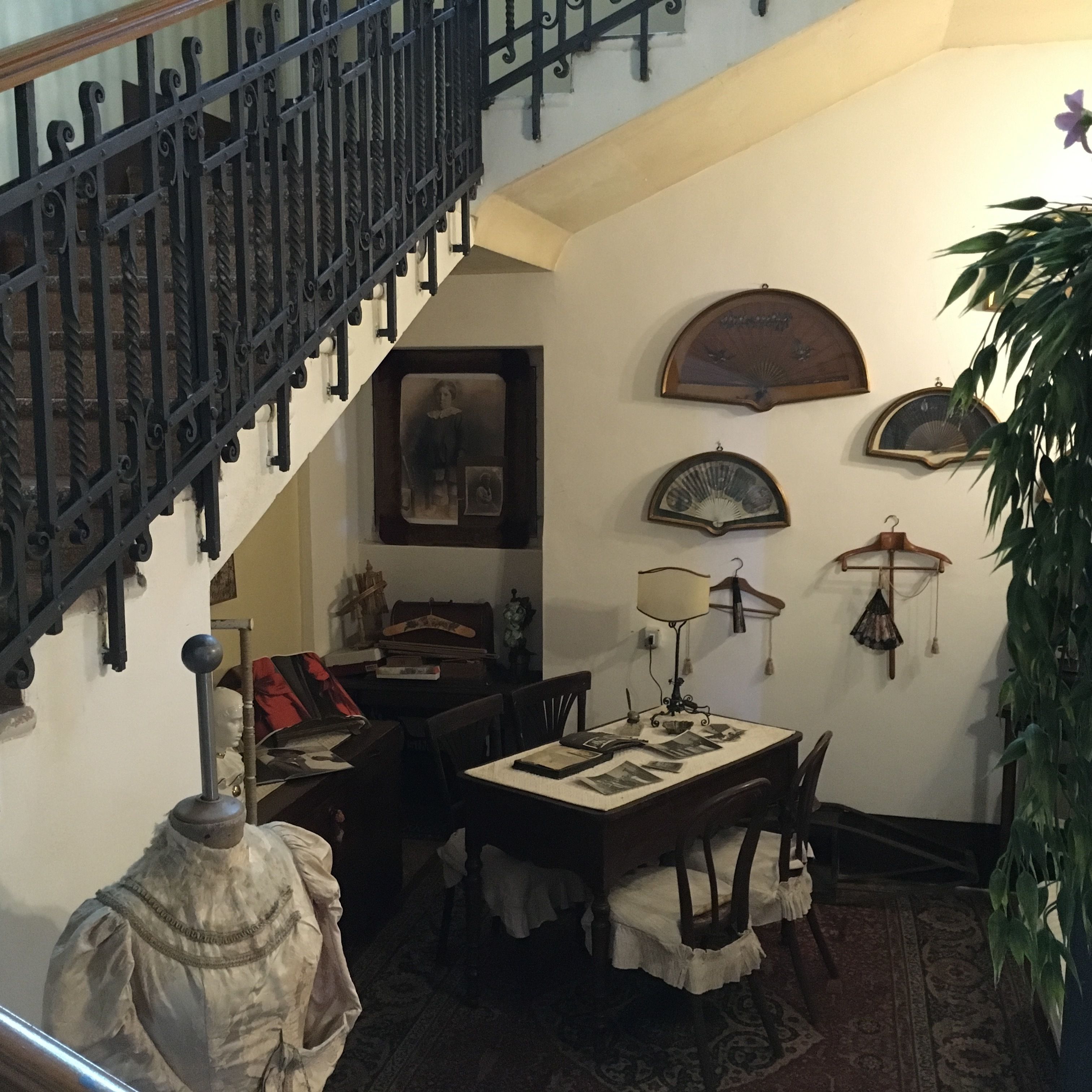 Uma escadaria com grades pretas ricamente decoradas, uma área de refeições com uma mesa e cadeiras, e uma parede com quadros e um relógio.