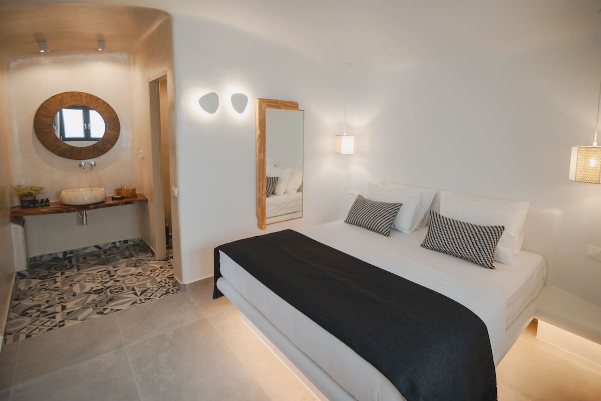A modern bedroom with a large bed, a round mirror, a bedside table, and a bathroom with a sink and patterned floor.