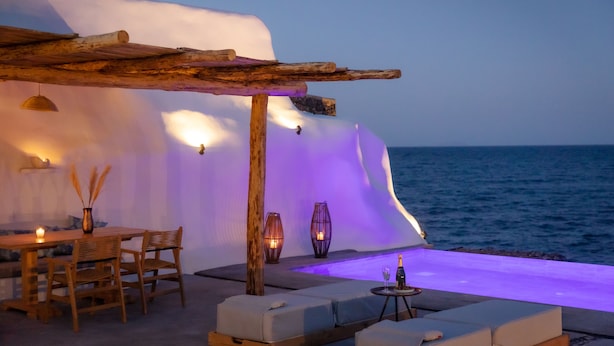 Eine Strandlounge mit Holztisch, gepolsterten Sitzmöglichkeiten und stimmungsvoller Beleuchtung.