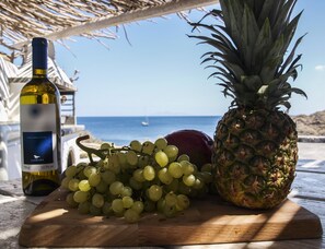 A wooden cutting board with a bottle of wine, grapes, and a pineapple on a terrace with a view of the sea. - Casa Di Mare (Santorini)