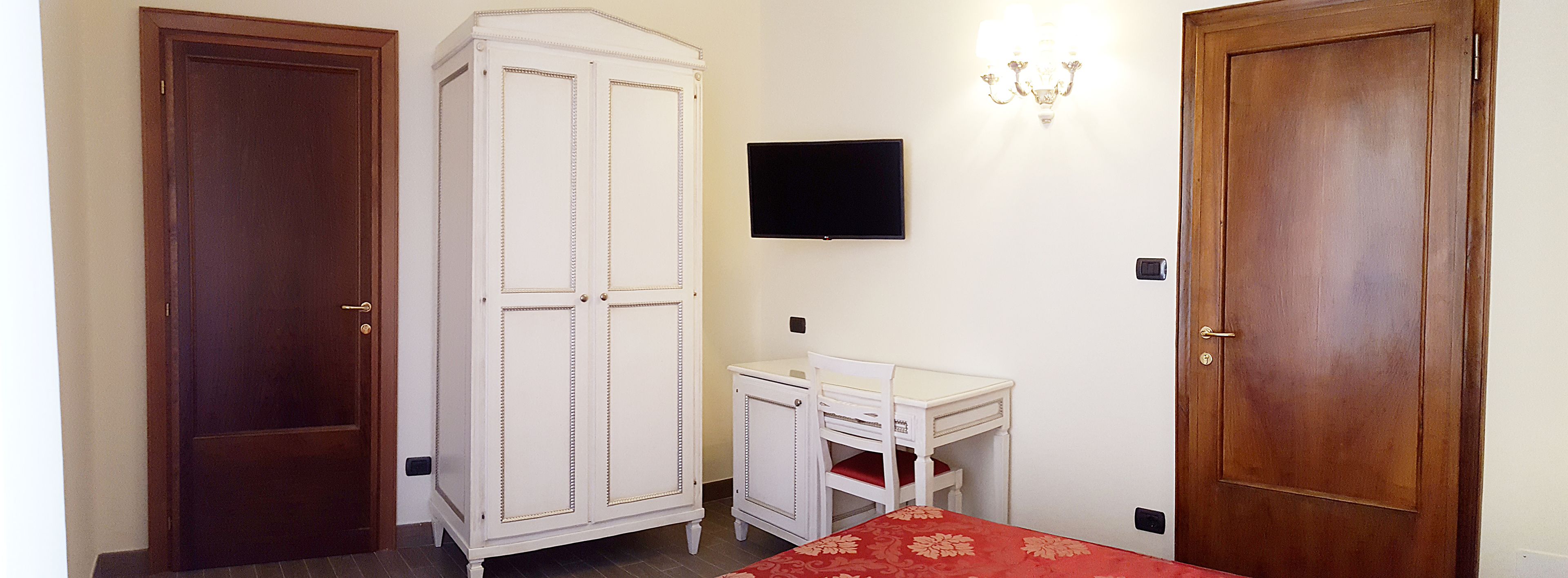 A bedroom with two doors, a wardrobe, a desk, a chair, a television, and a chandelier.
