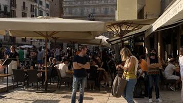 An outdoor cafe area with tables and umbrellas, surrounded by buildings with balconies and a tower in the background.