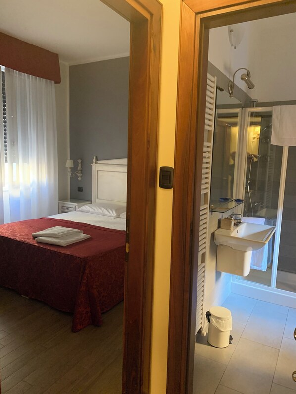 Una habitación de hotel con cama, mesita de noche, baño con lavamanos y espejo, y ducha.