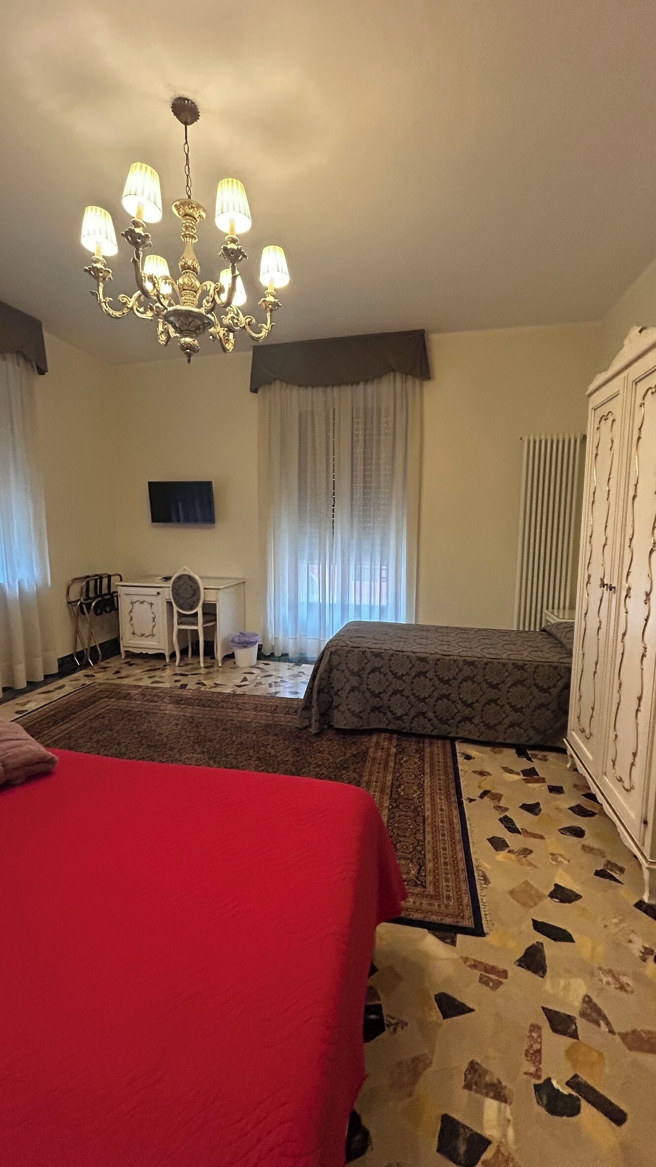 A hotel room with a large bed, a smaller bed, a desk with a chair, a chandelier, and a radiator.
