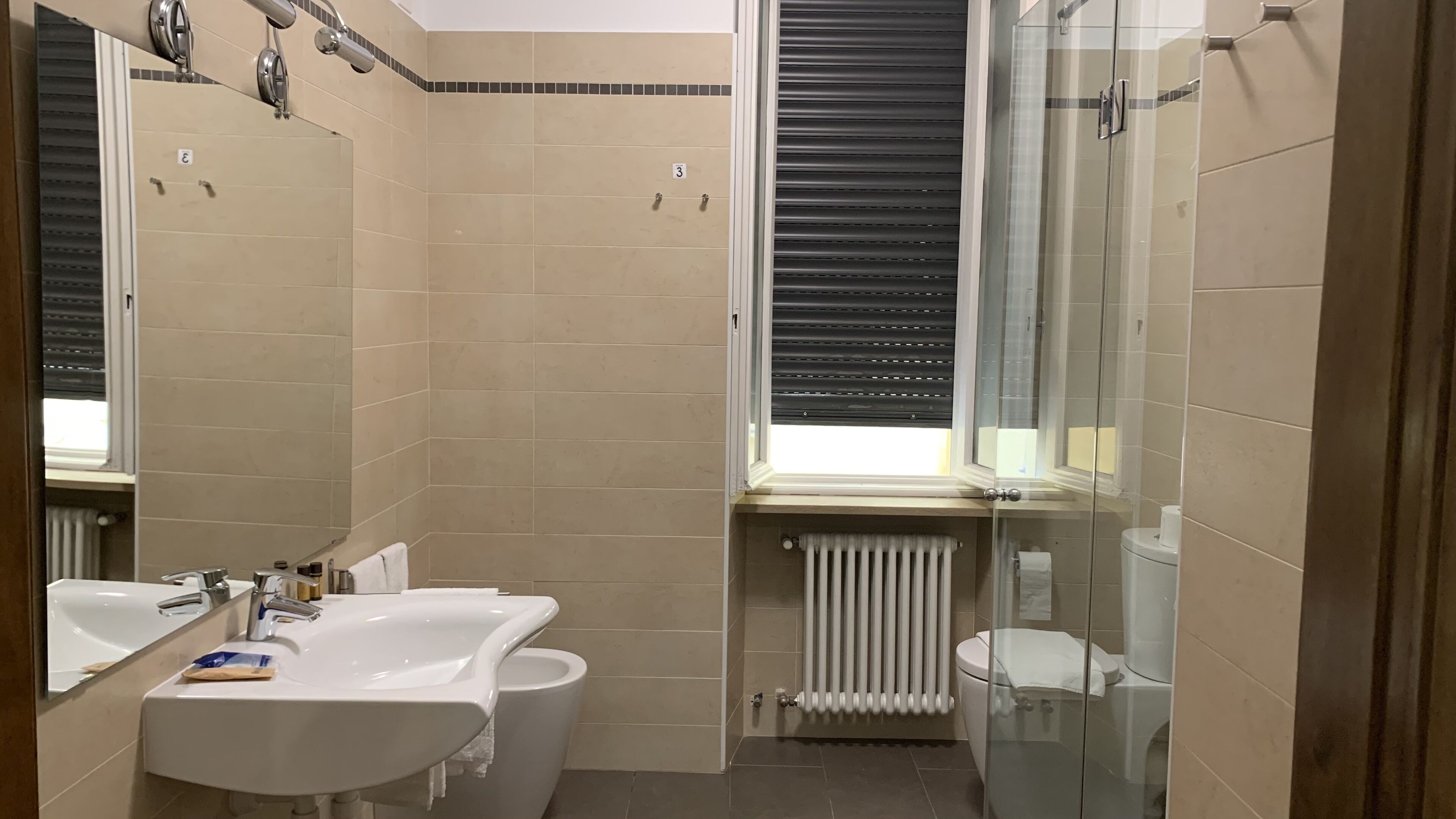 Een badkamer met twee wastafels, een toilet, een douchecabine van glas en een raam met zonwering.